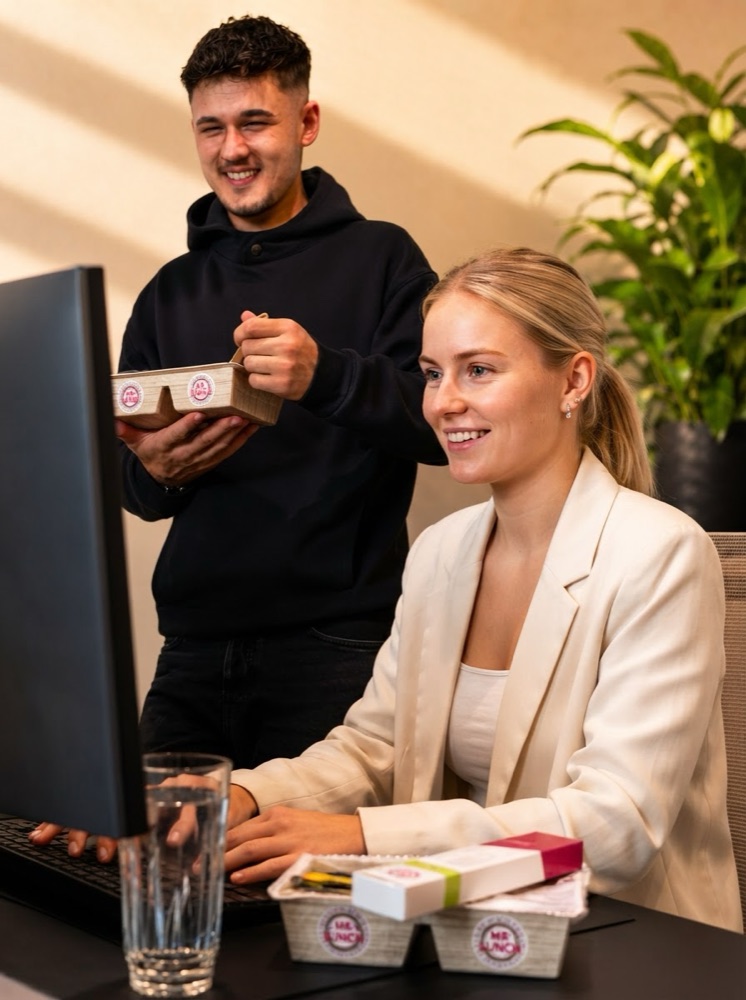 Ein Kollege hält eine Mr.-Lunch-Menüschale, eine Kollegin am Schreibtisch lächelt auf den Bildschirm